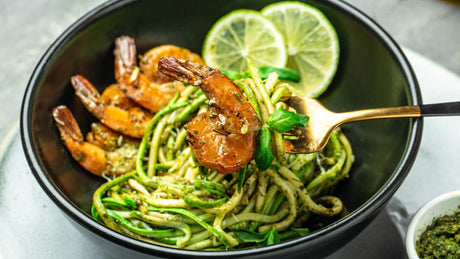 Courgettepasta met garnalen, pesto en limoenschijfjes in zwarte kom.
