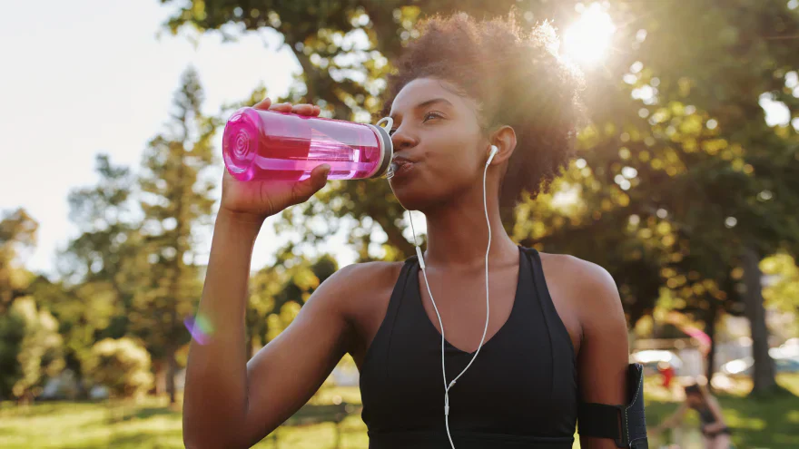 Femme qui boit de l'eau dans une gourde rose en extérieur après une course, avec des écouteurs et le soleil filtrant à travers les arbres