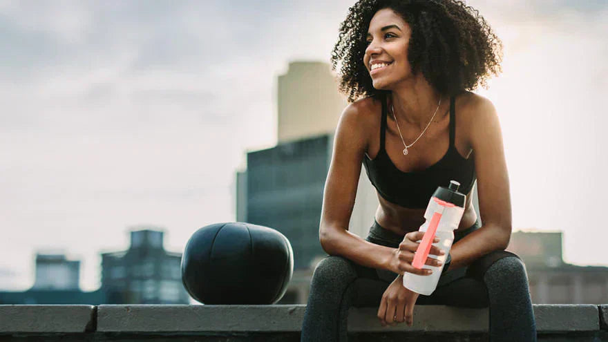 Woman sitting outdoors after a workout holding a water bottle, with a medicine ball beside her