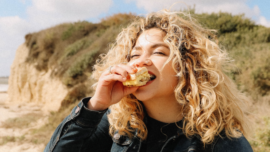 Femme aux cheveux bouclés mangeant un sandwich sur la plage.