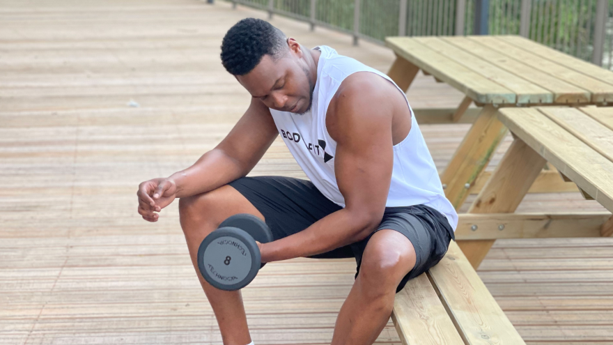 Man doet bicep curls met dumbbell op houten bankje buiten.