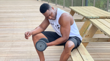 Man doet bicep curls met dumbbell op houten bankje buiten.