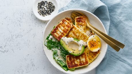 Grilled halloumi, avocado, egg, and greens in white bowl.