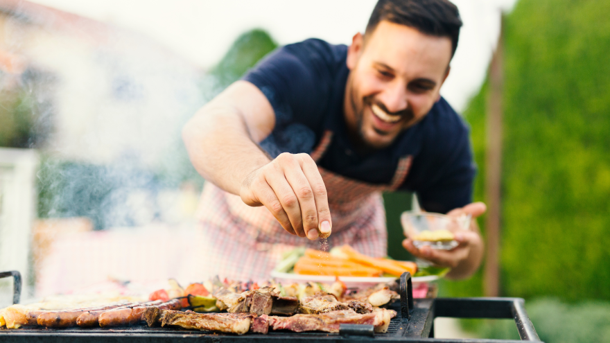 Man kruidt vlees op barbecue met groenten op achtergrond.