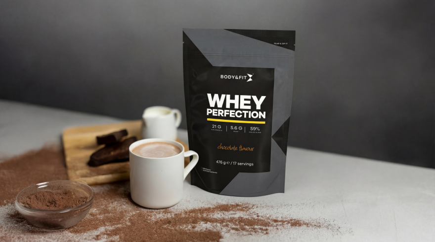 Body&Fit whey protein next to a cup of hot chocolate with cocoa powder and chocolate pieces