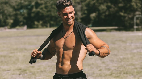 Shirtless man outdoors smiling and holding shirt over shoulders.