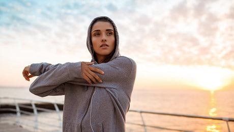 Woman in hoodie stretching outdoors at sunrise.